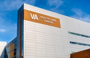 Va Health Care Center's Exterior Facade Brand And Logo Signage On A Bright Sunny Day With Blue Sky, Hazy Clouds, Floors And Windows.